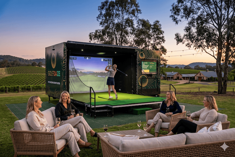 Group of women relaxing with wine on lounges while a friend plays the Golf Daze simulator at a private vineyard hens party